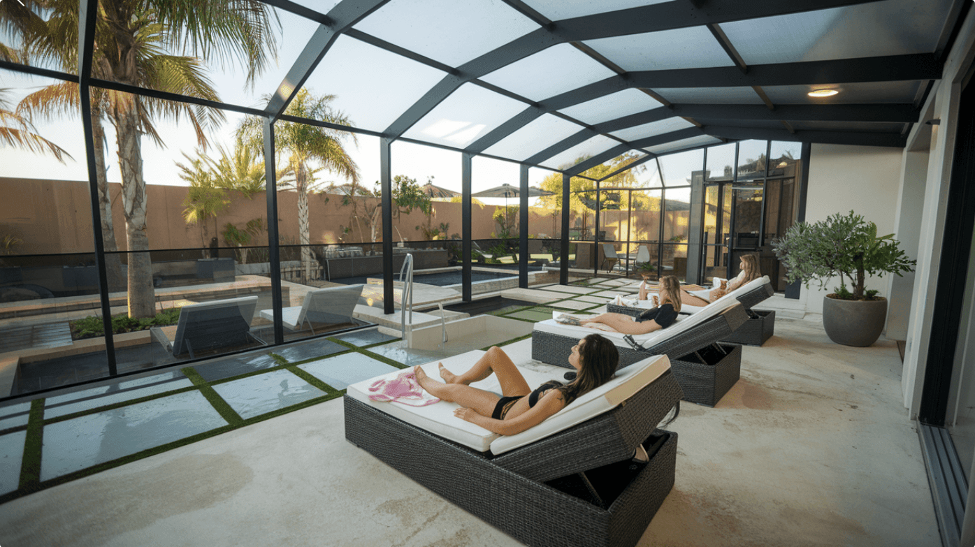 People relaxing on lounge chairs under a screened patio with a pool and palm trees in the background.